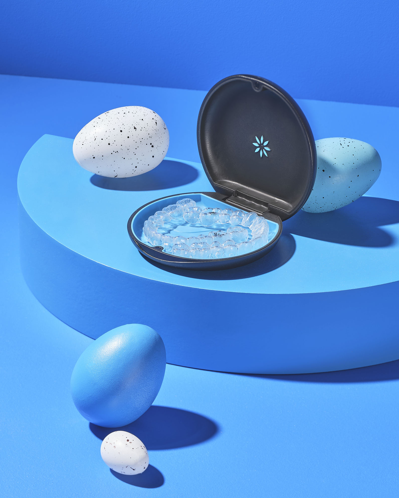 Clear aligner in a black case surrounded by decorative eggs on a blue background.