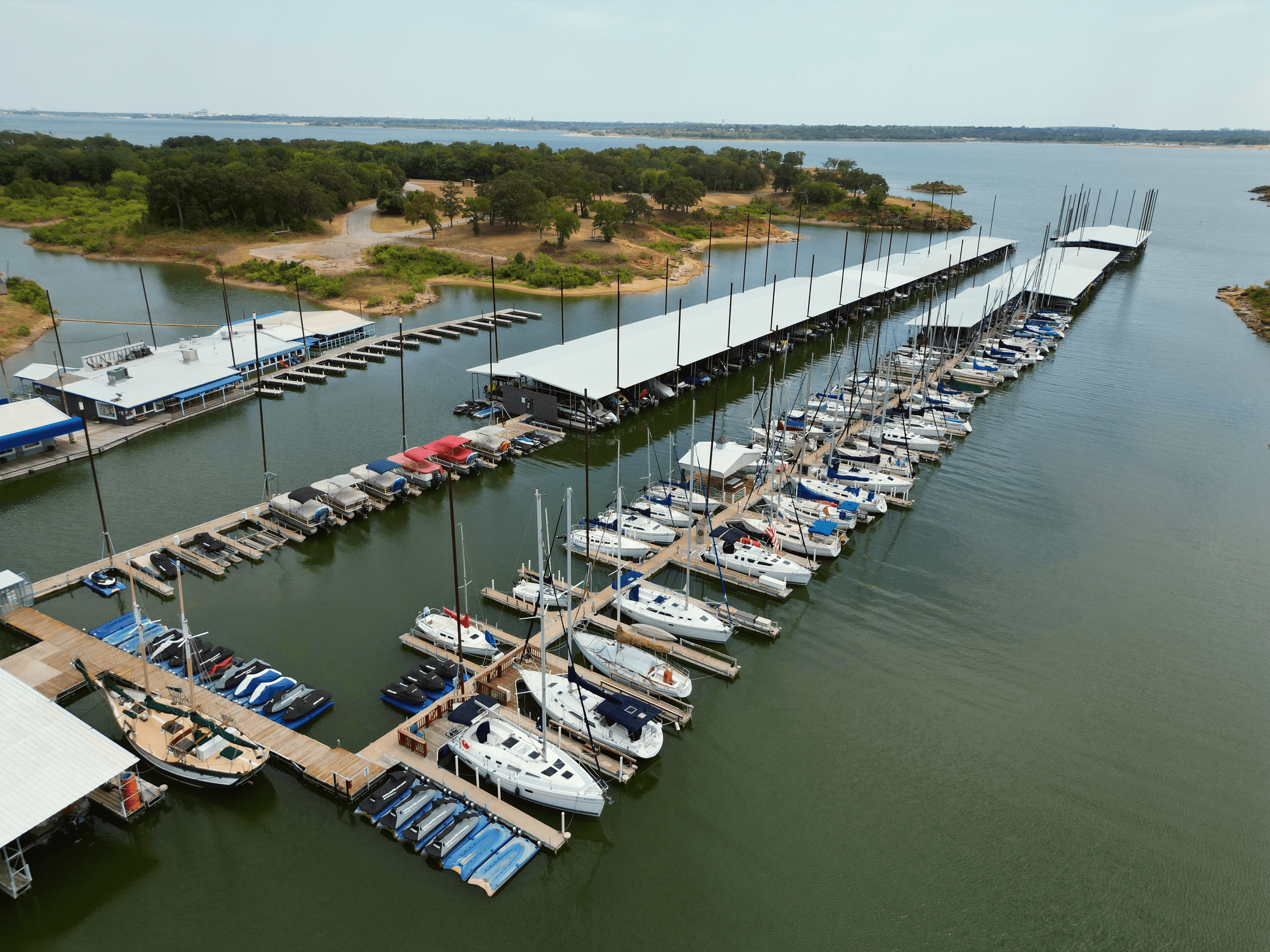 Aerial view of a marina with numerous sailboats and yachts docked along multiple piers, surrounded by calm water and lush greenery in the background.