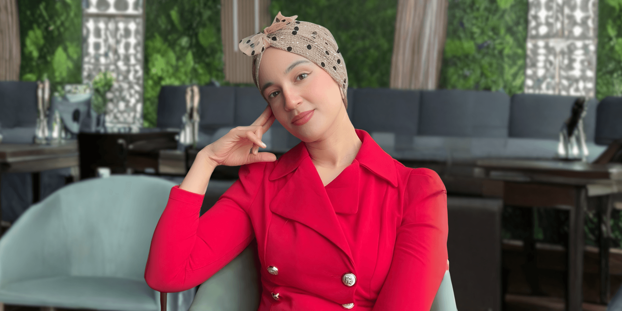 Woman in red blazer and polka-dot headscarf sitting thoughtfully in a modern café setting.