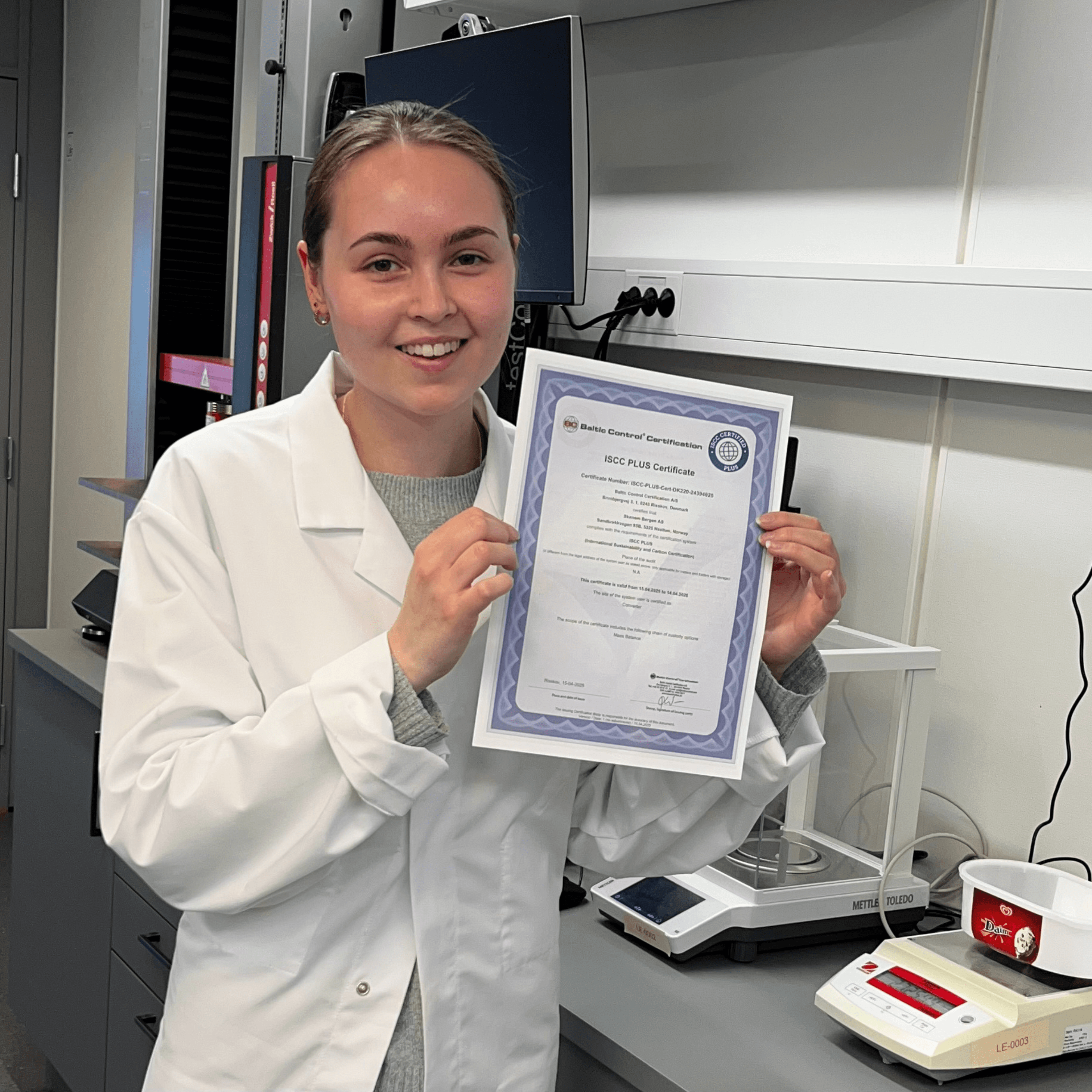 Young woman in labcoat smiling, holding up a certificate