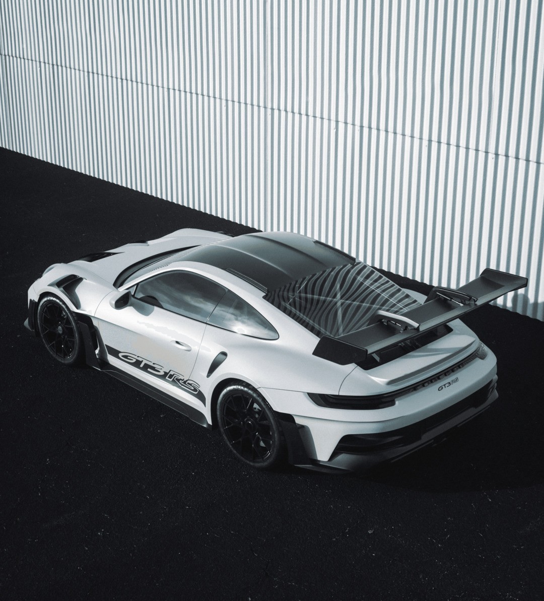 A sleek, white sports car with aerodynamic features and a large rear spoiler is parked on a dark asphalt surface next to a striped, industrial wall, highlighting its sporty and modern design.
