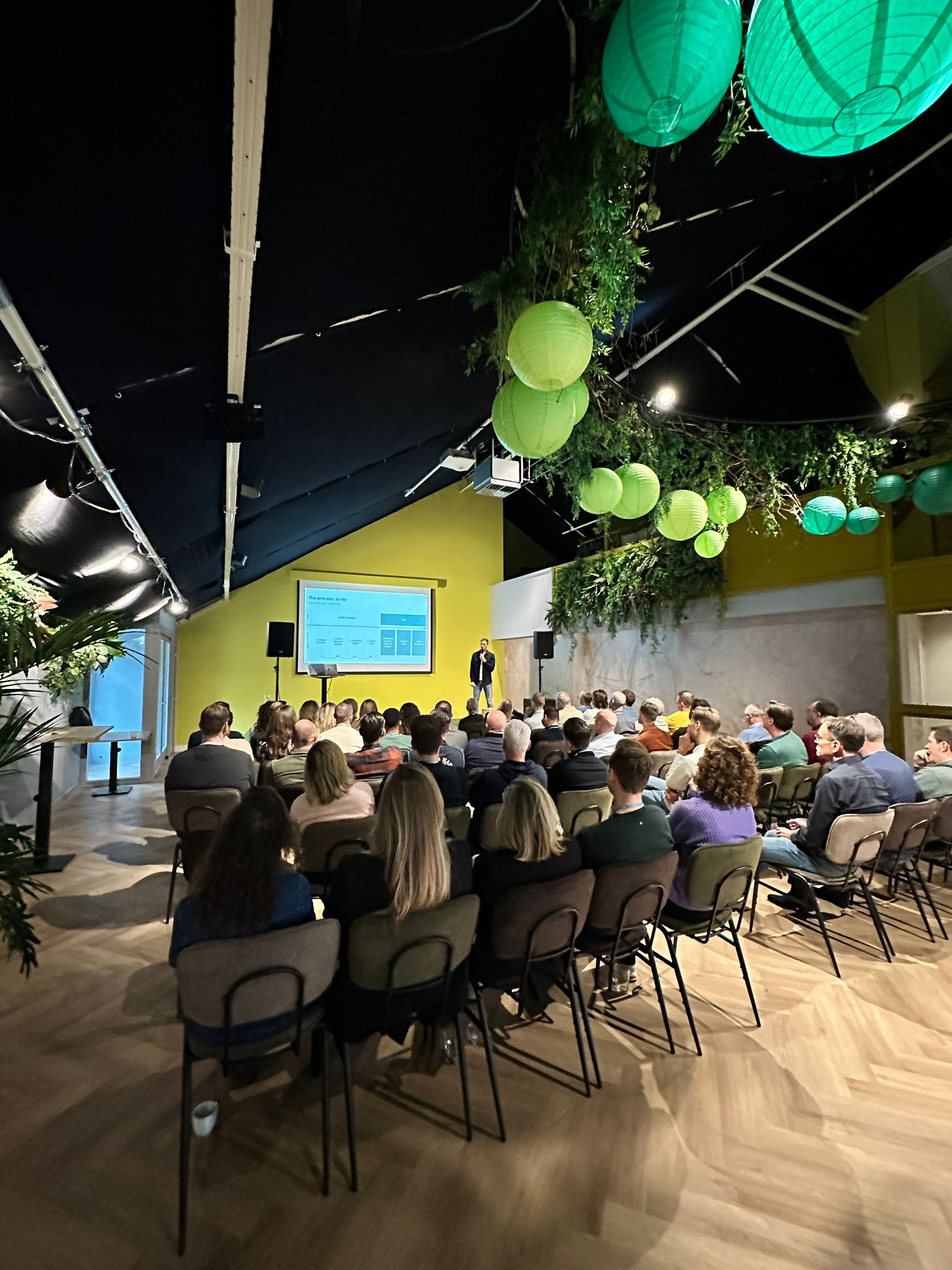 A crowd of people seated in a conference room, with vibrant green decor and a presentation screen at the front.