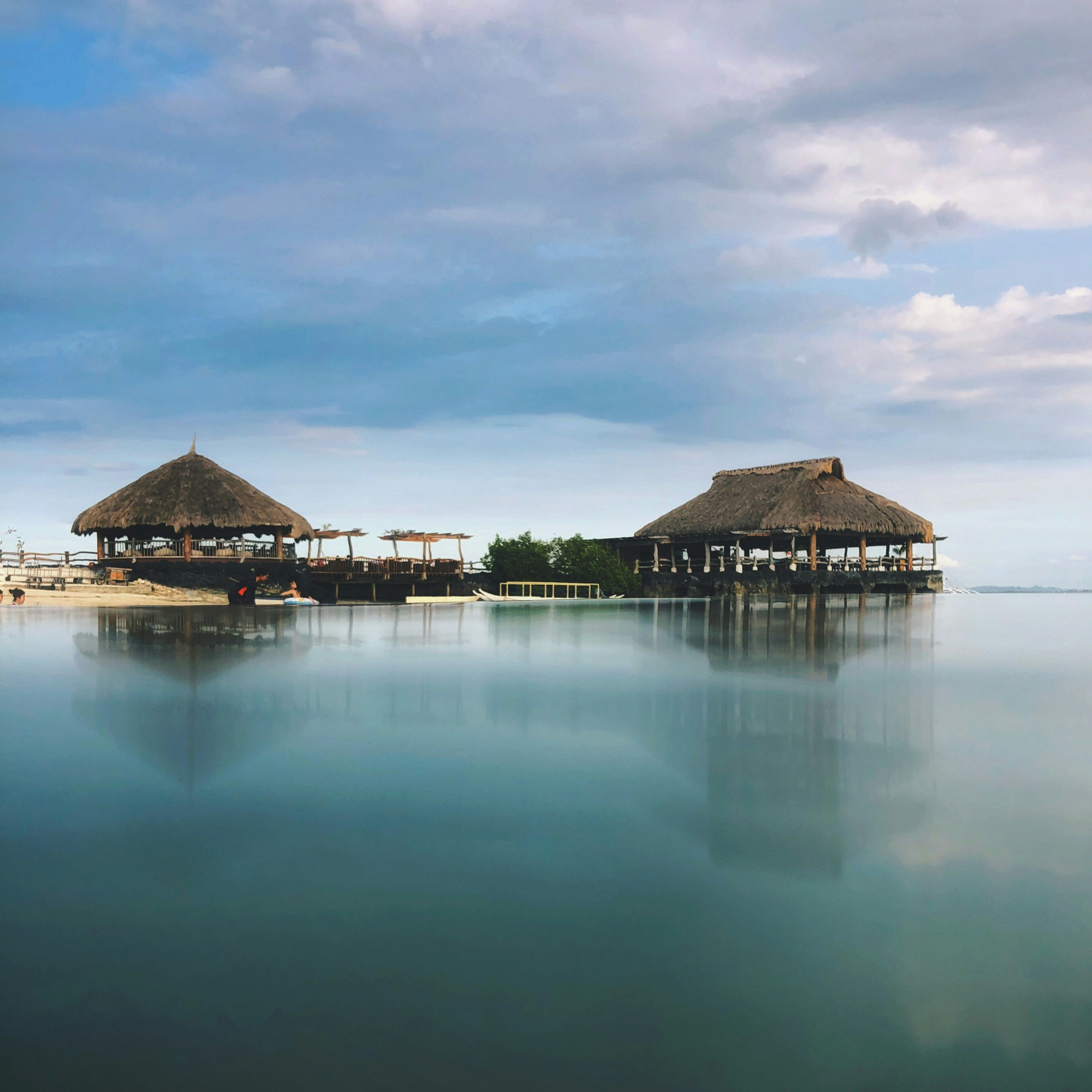 Two thatched-roof huts sit over calm waters under a serene sky, reflecting tranquility and nature's beauty.