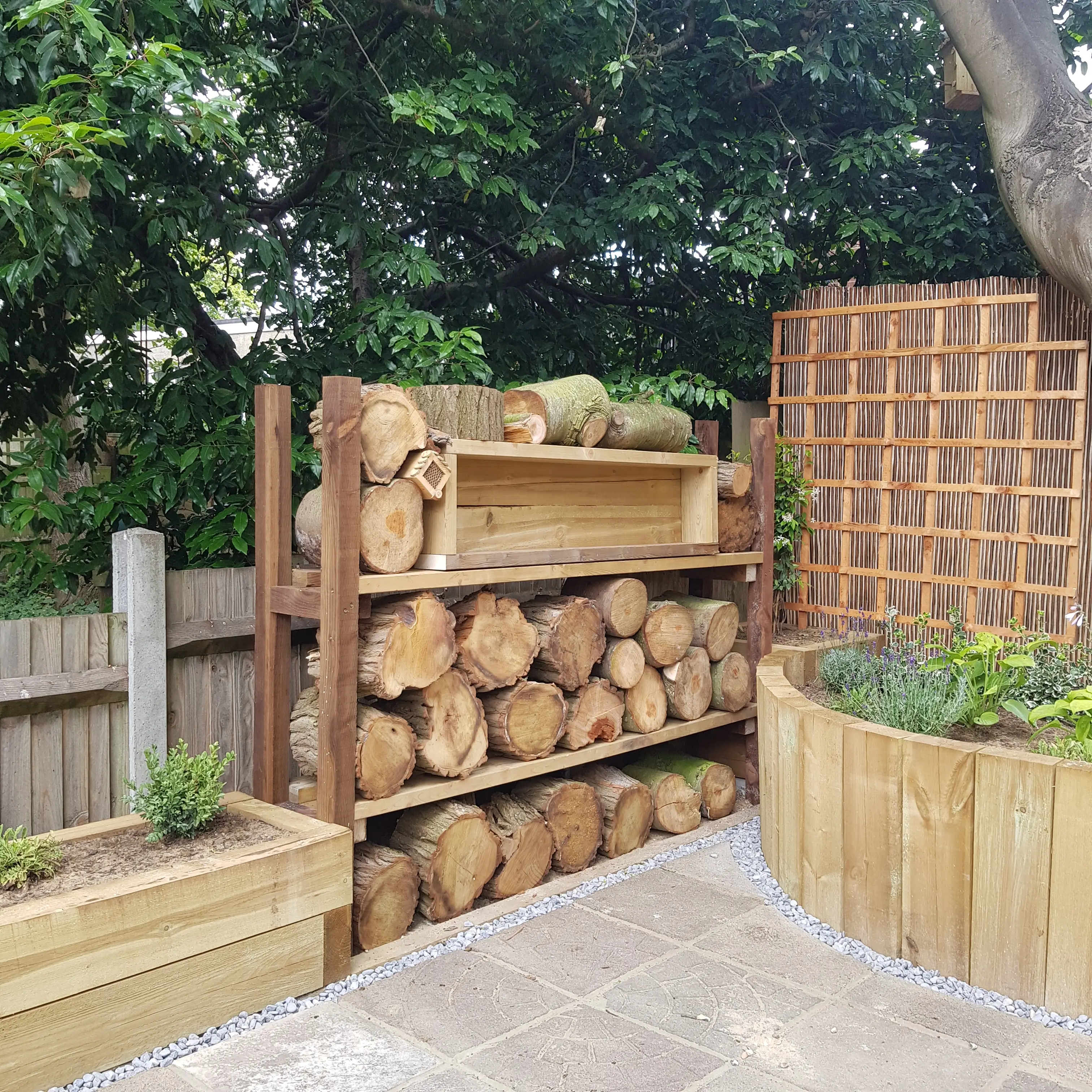 A wooden outdoor storage area displays stacked logs beside a raised garden bed and a wooden fence.