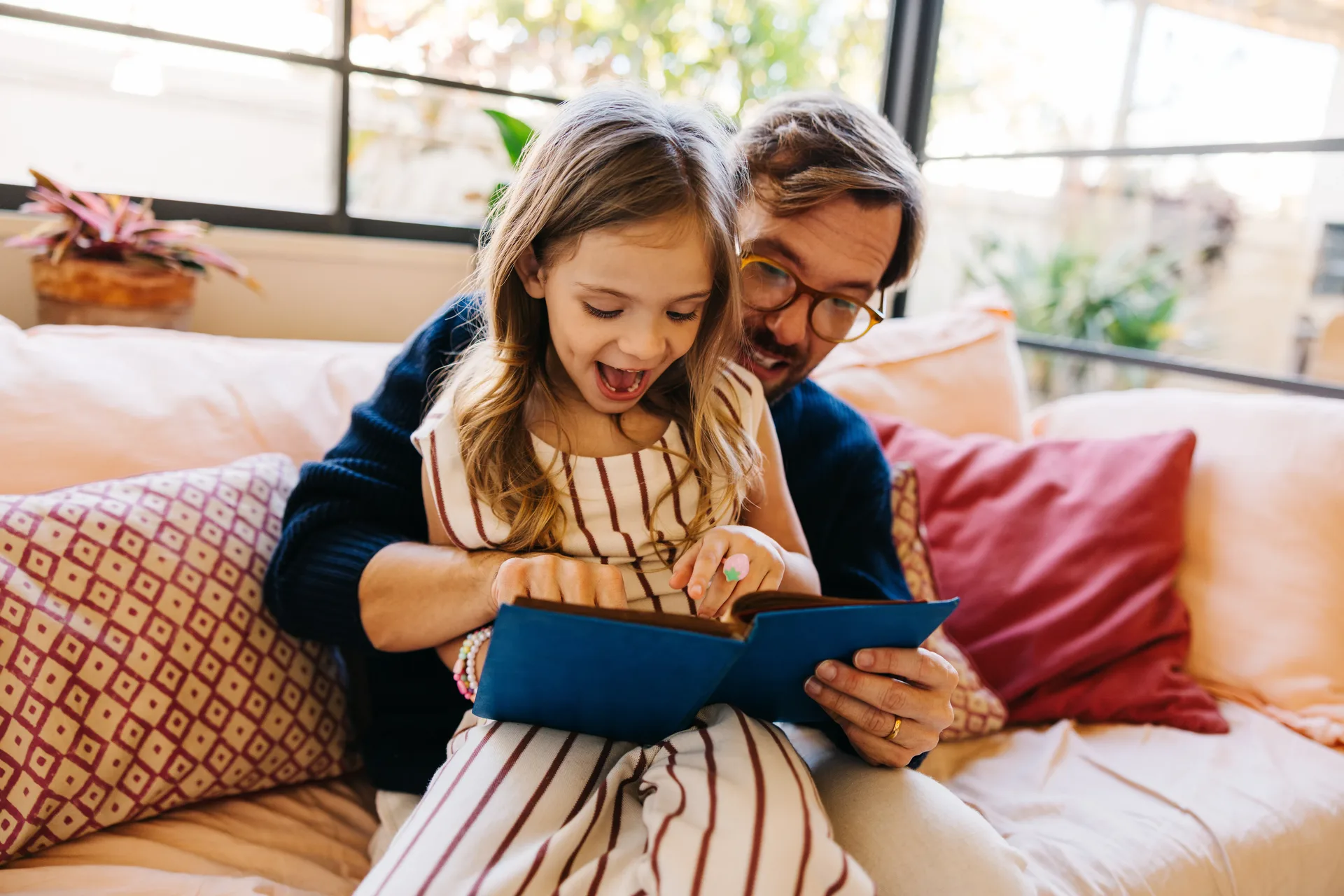 A father and young daughter sitting on a couch together, laughing as they read a book