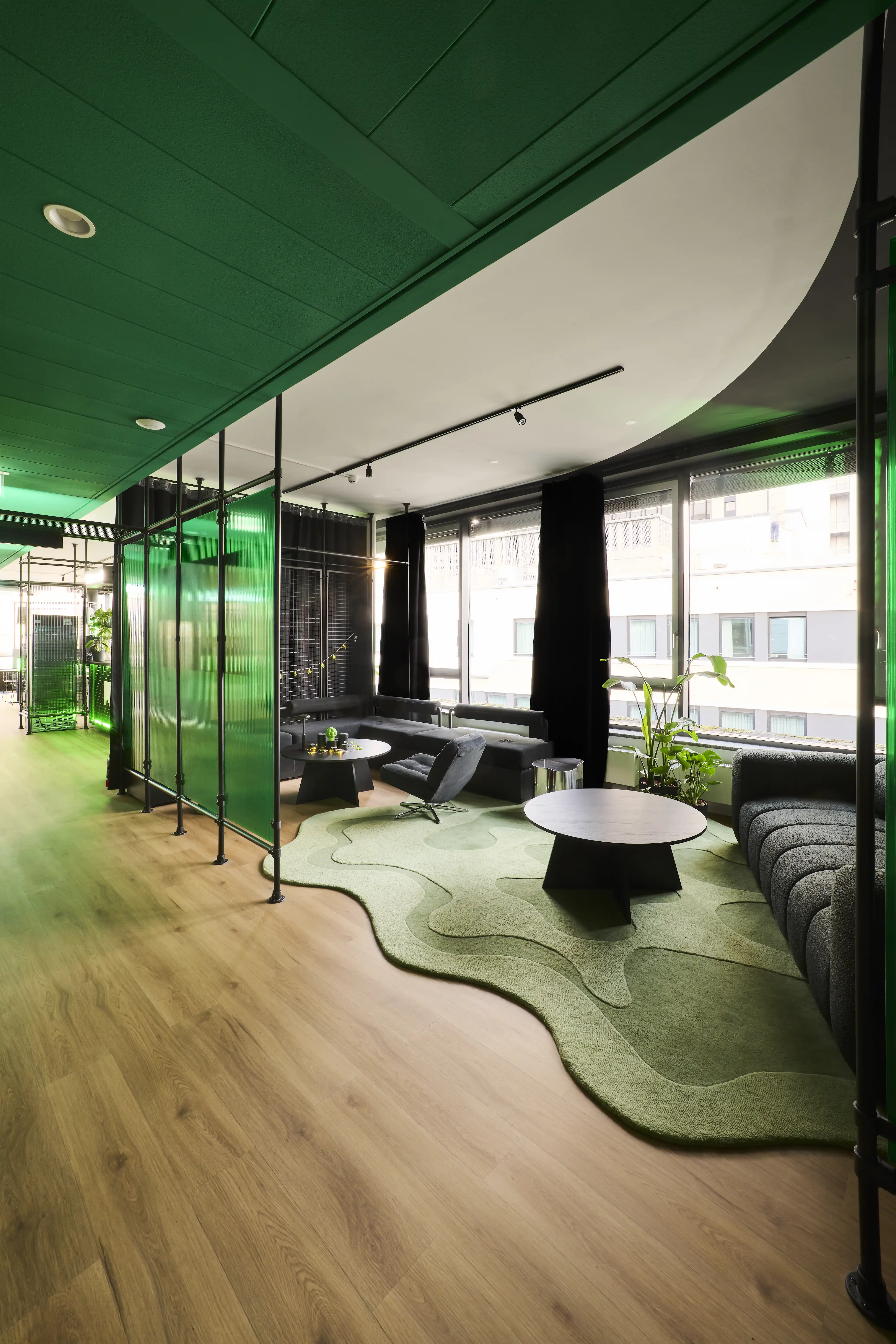 Window lounge at BLOOMWELL with black upholstered sofas, round black coffee tables, an organically shaped green rug, green glass partitions and a sweeping green-and-white ceiling.
