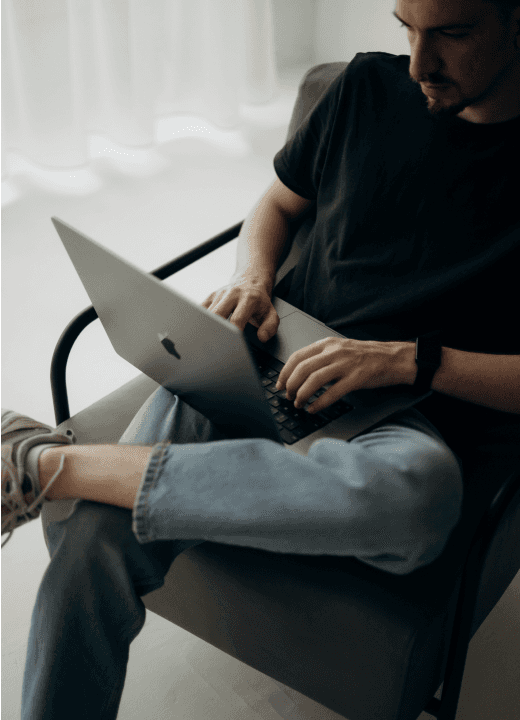 A person wearing a black t-shirt and jeans sits in a modern armchair, focused on typing on a sleek, open laptop in a bright room with sheer curtains.