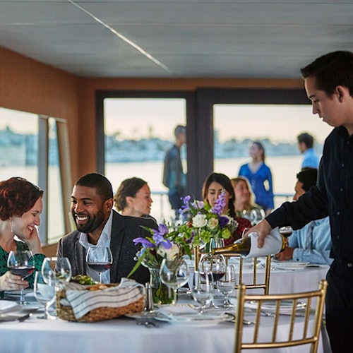 A group of people dine at an elegantly set table on a boat. A waiter pours a drink while others chat and smile in the background.
