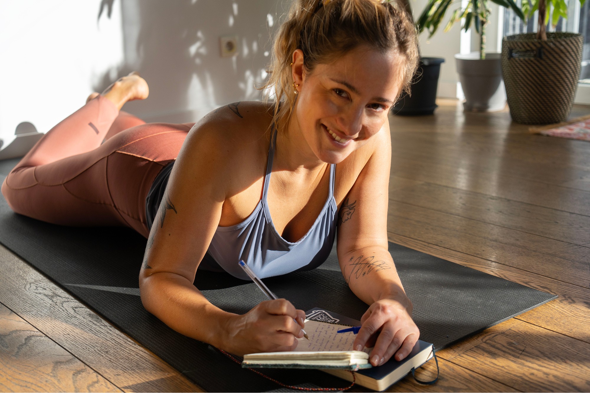 A woman lies on a yoga mat, smiling and writing in a journal, with sunlight streaming through a window, creating a peaceful atmosphere for mindfulness and relaxation.