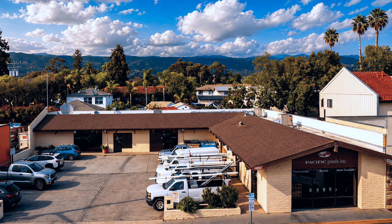 Wider aerial view of the same single-story commercial property showing a flat-roofed retail or service building with a full parking lot of work trucks and service vehicles, a tenant signage panel visible at the corner, and mountain and tree views in the background under a partly cloudy sky.