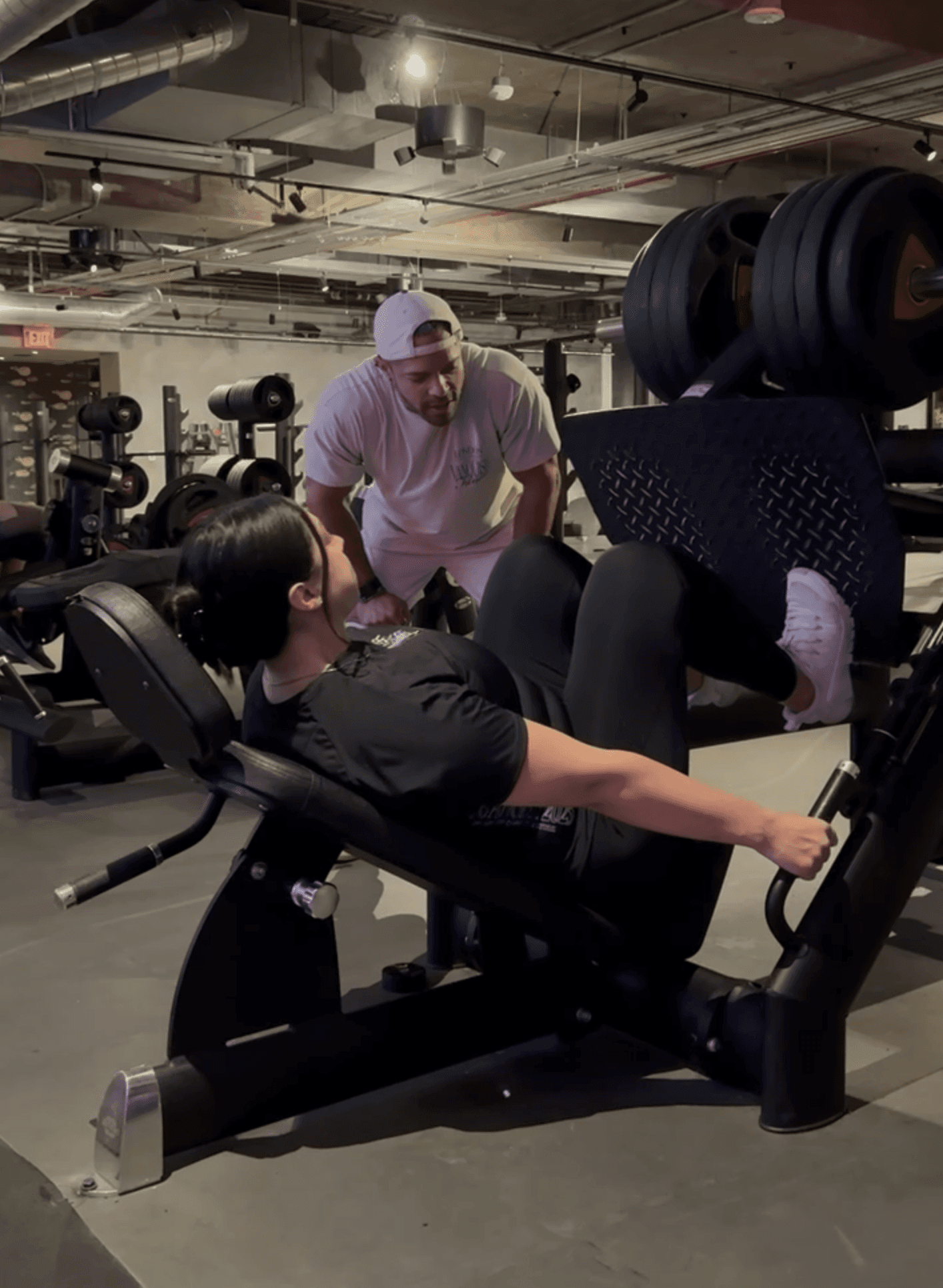 A muscular man smiling while coaching a lady on the leg press machine