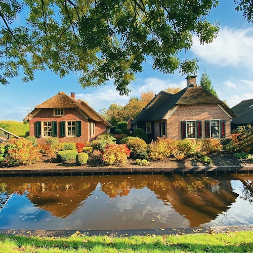 Dua pondok beratap rumbai dengan taman yang berwarna-warni terletak di samping kanal yang tenang dan reflektif di bawah langit biru dengan awan yang tersebar.