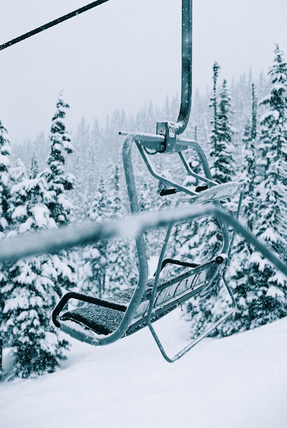 A snow-covered chairlift moves through falling snow past a pine forest
