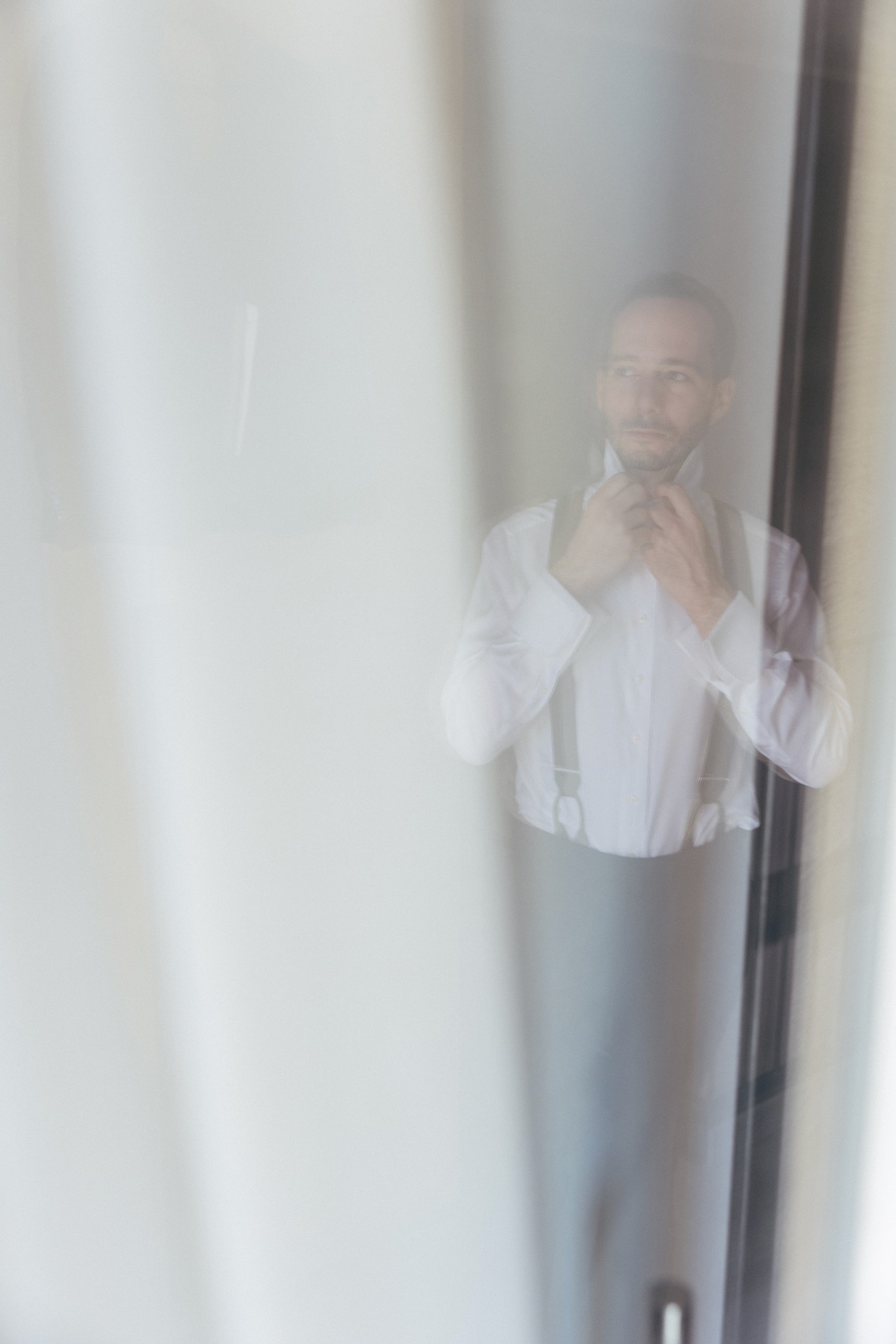 Sposo in camicia bianca e gilet ai preparativi matrimoniali, riflesso in vetro con luce naturale diffusa