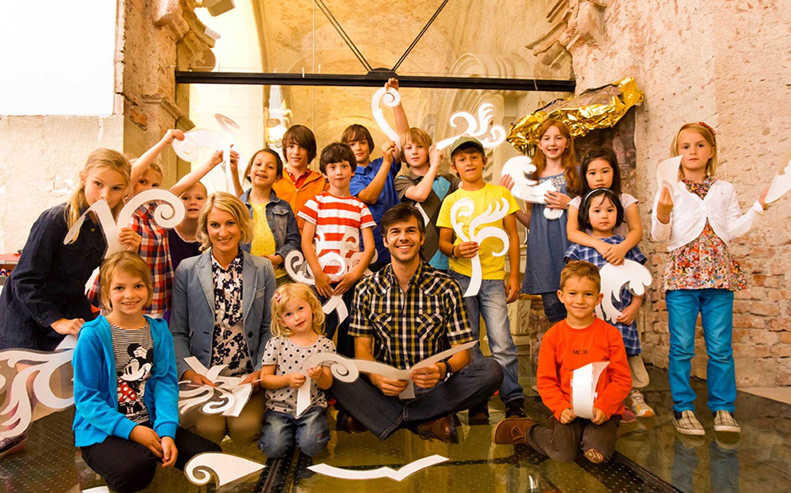 Children and adults holding decorative paper cutouts inside Klosterneuburg Monastery.