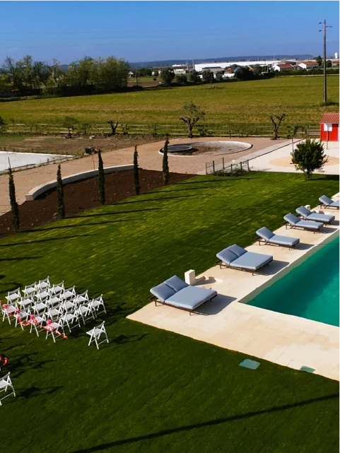 A serene poolside scene with six gray lounge chairs on a stone patio, next to a turquoise pool. Rows of white chairs on a lush green lawn face a field.