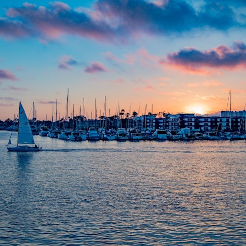 Sailboat on a calm waterway with numerous docked boats and buildings in the background under a vibrant sunset sky.