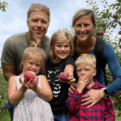 Krista Lehde smiling outdoors with her family in an apple orchard, with children holding freshly picked apples.