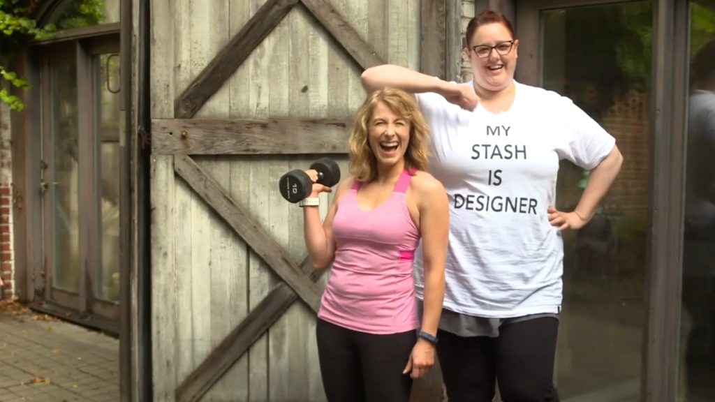 Two people stand happily outside a wooden barn-style building; one is wearing a pink athletic top and holding a dumbbell, and the other sports a white t-shirt