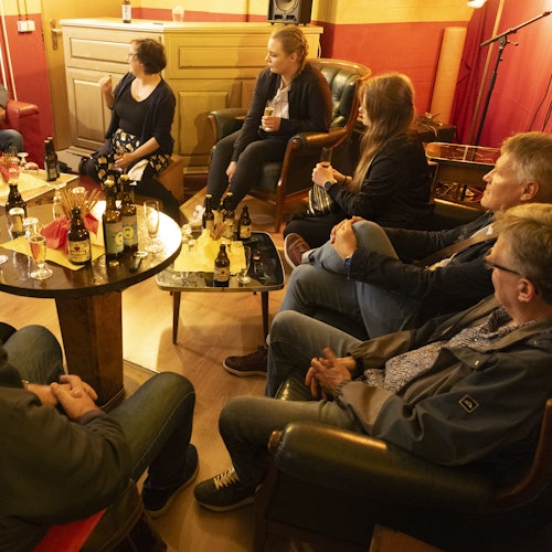 People sitting in a cozy room, chatting and drinking, with bottles and glasses on tables.