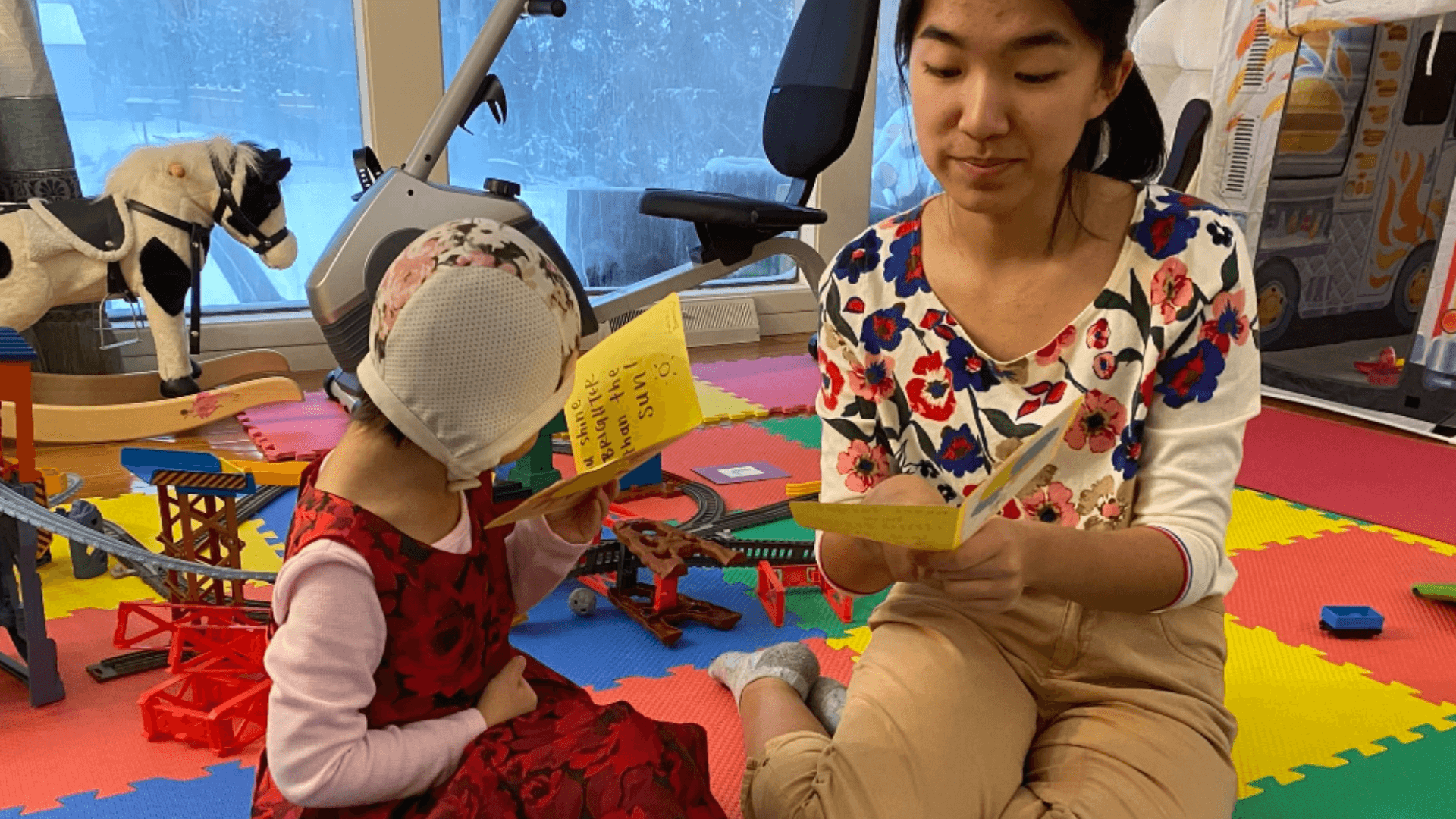 older teen and young child sit on colorful foam mats reading yellow picture books togehter, surrounded by toy train tracks and play equipment in an indoor playroom