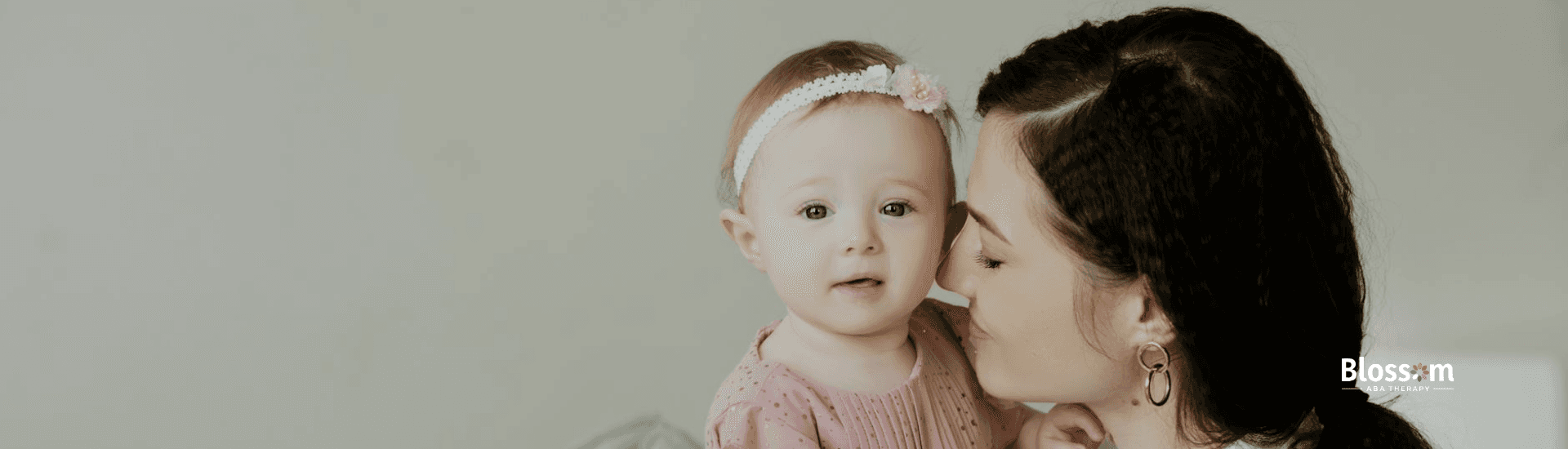 A mother lovingly holding and kissing her autistic baby, who is wearing a pink dress and headband.