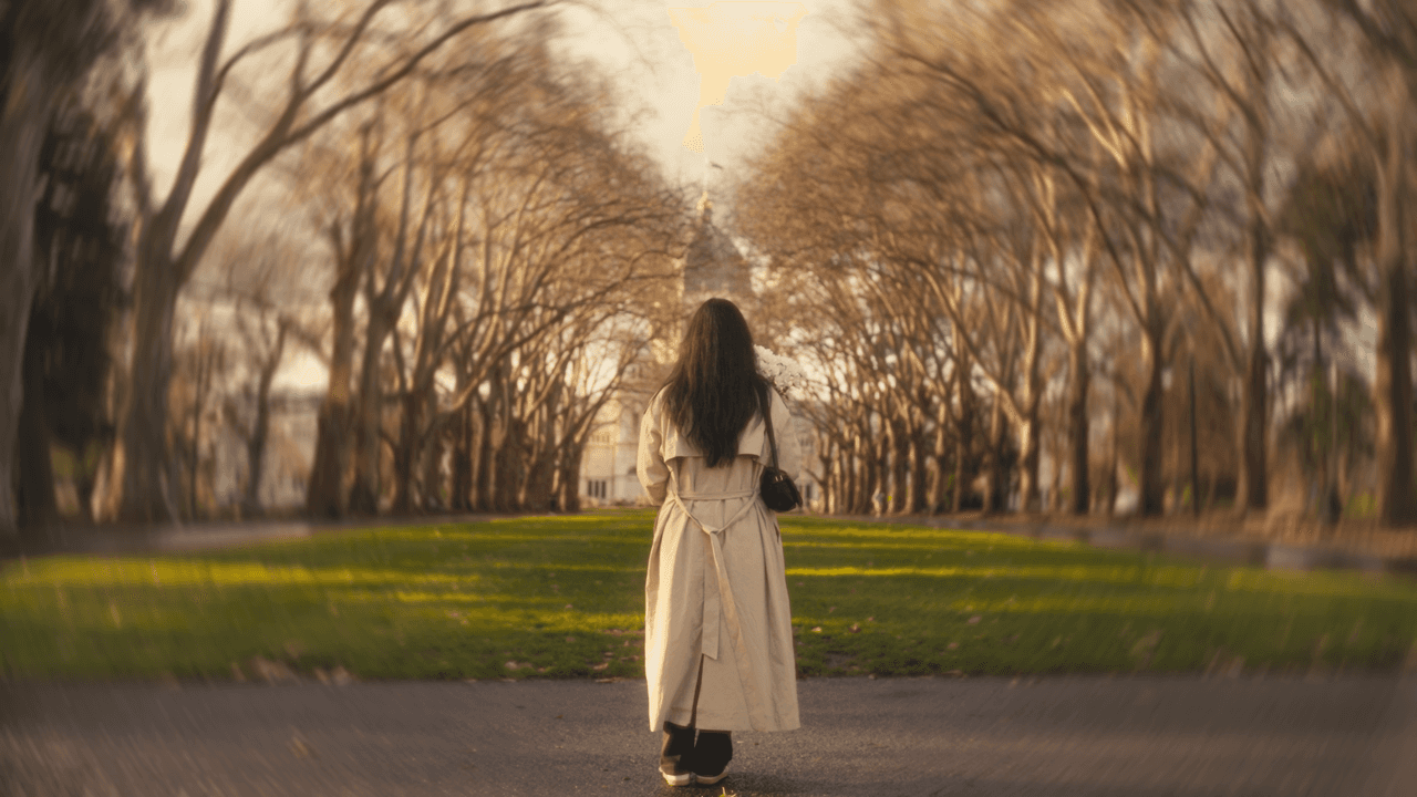 A back shot of a woman wearing a long coat, walking in an autumn park, with barren trees and soft light filtering through