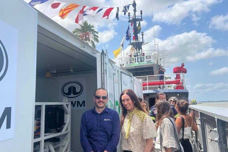 Homem de camisa azul e mulher de blusa clara com estampa estão em primeiro plano em embarcação. Ao fundo, outras pessoas conversam e há bandeiras náuticas coloridas penduradas acima. Céu parcialmente nublado e estrutura de navio visível.