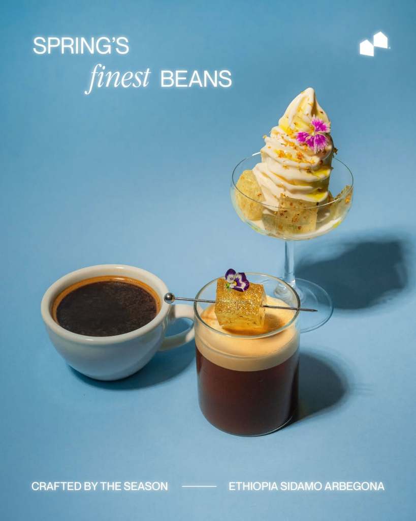 A display of coffee and dessert featuring a cup of dark coffee, a glass of layered coffee topped with cream and decorative flower, and a dessert of soft serve ice cream garnished with petals on a blue background.