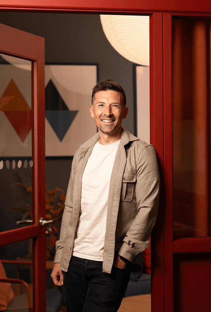 Photo of Harry Armstrong smiling at the camera in an open doorway wearing a beige open shirt and a white t-shirt