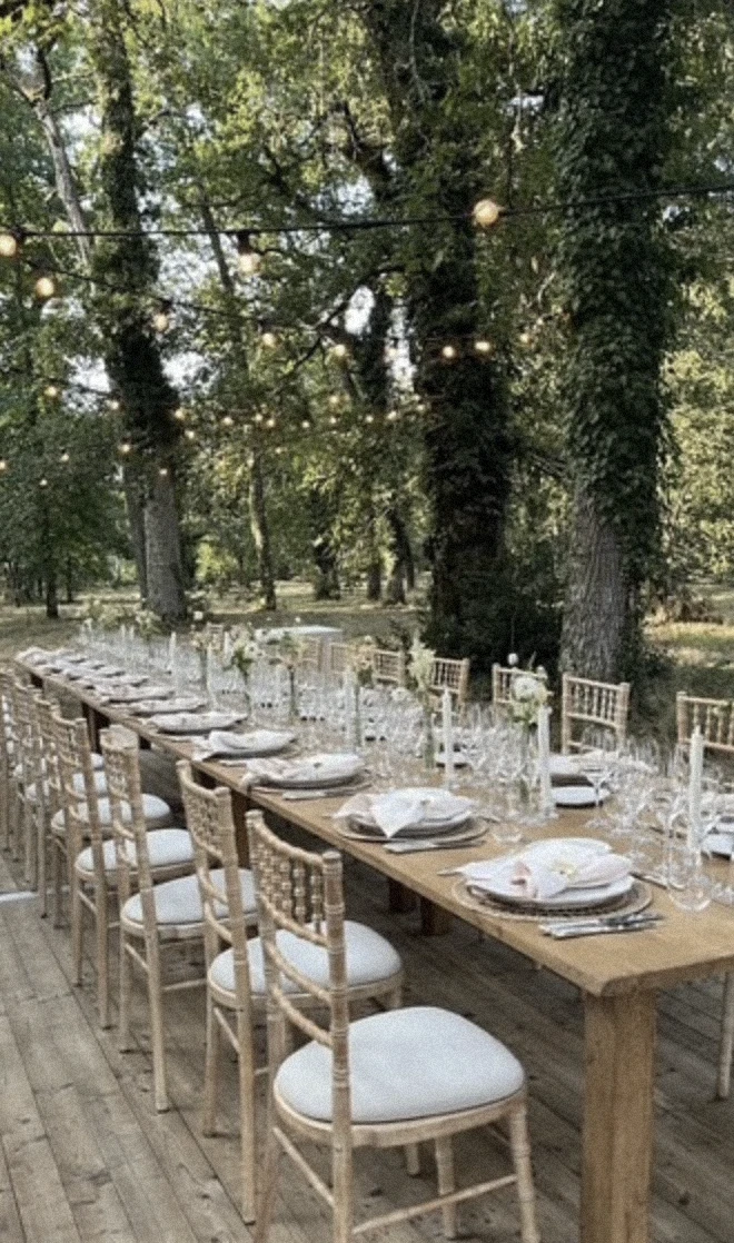 A long outdoor dining table surrounded by trees and string lights, set for an intimate evening gathering in a romantic woodland setting.  