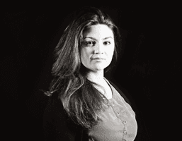 A black and white portrait of a woman with long, dark hair. She is looking directly at the camera with a neutral expression. She is wearing a dark-colored top with buttons down the front. The background is solid black.