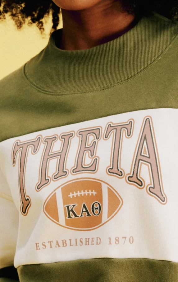 Close-up of a woman wearing an olive green mock-neck sweatshirt with a cream chest panel printed with "THETA."