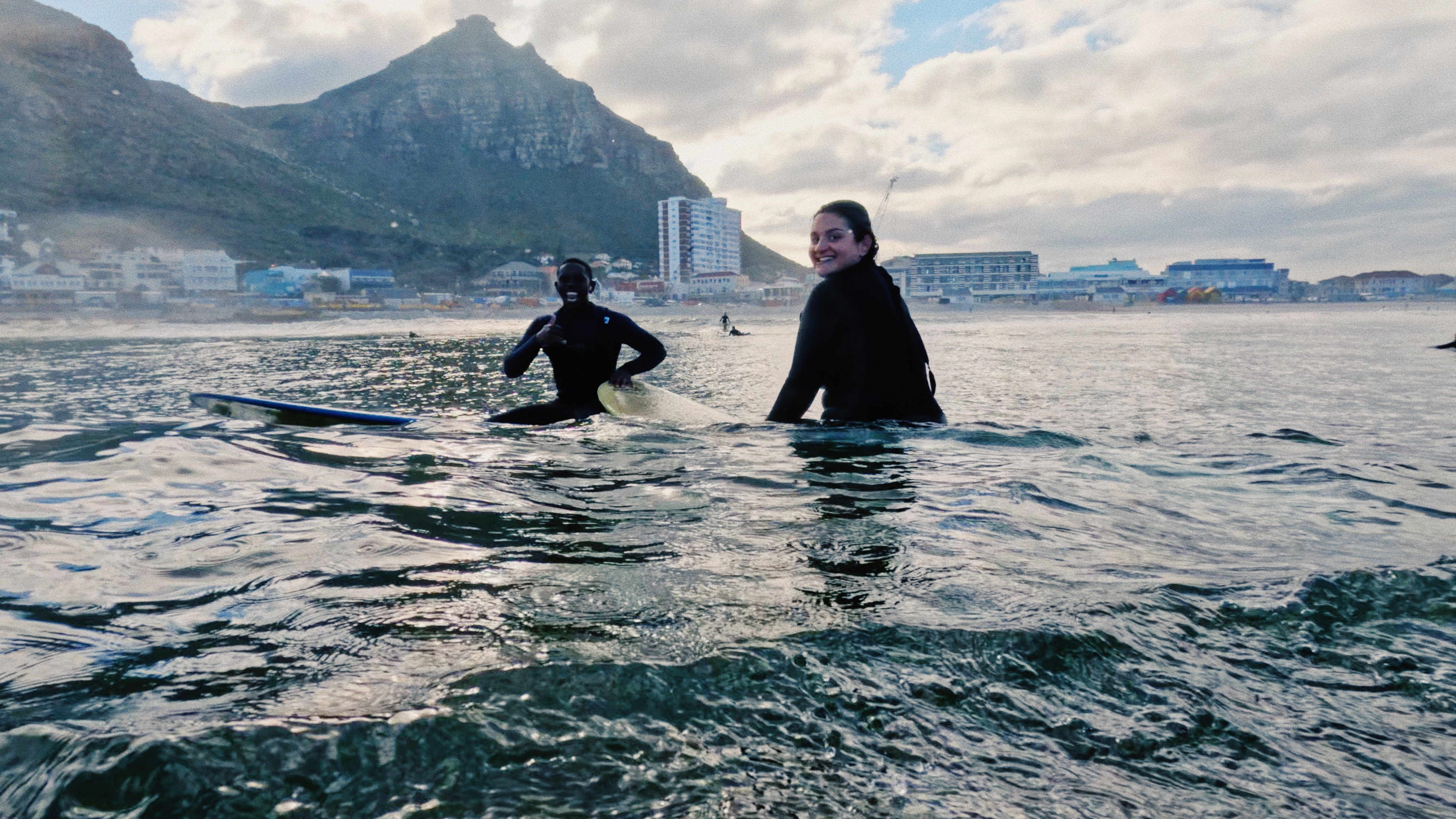 A surf coach is sitting on his board in the water, next to him is his female surf student sitting on her surfboard. The background shows beautiful Muizenberg in Cape Town
