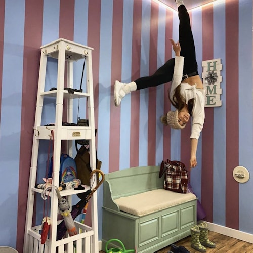 Person upside down on a striped wall, next to a green bench and a white shelving unit with various objects. Sign on wall reads "HOME."