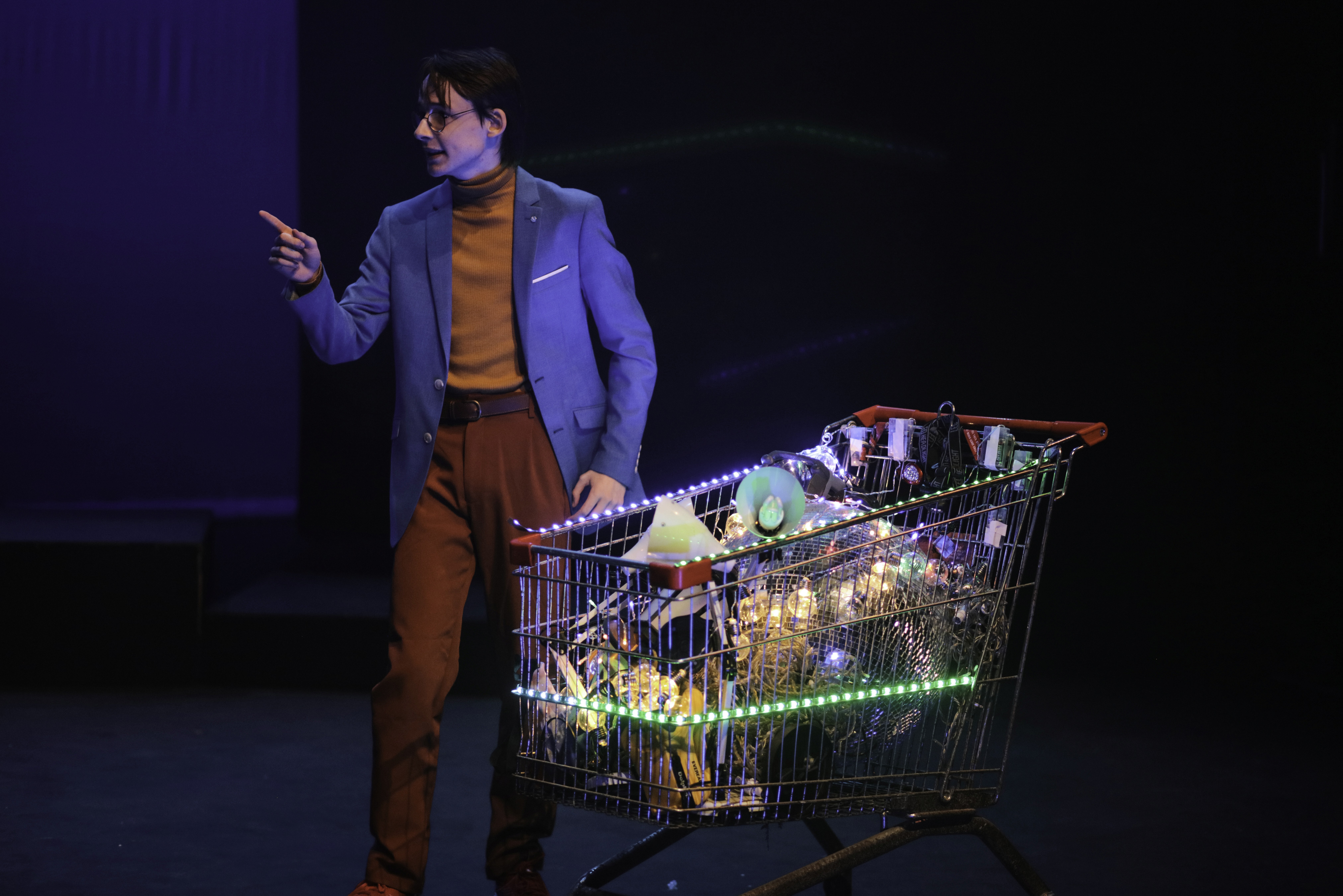 A performer dressed in a bright teal blazer and mustard turtleneck stands next to a shopping trolley filled with glowing lights and eclectic objects. They point outward, as if delivering a persuasive pitch under dramatic stage lighting.