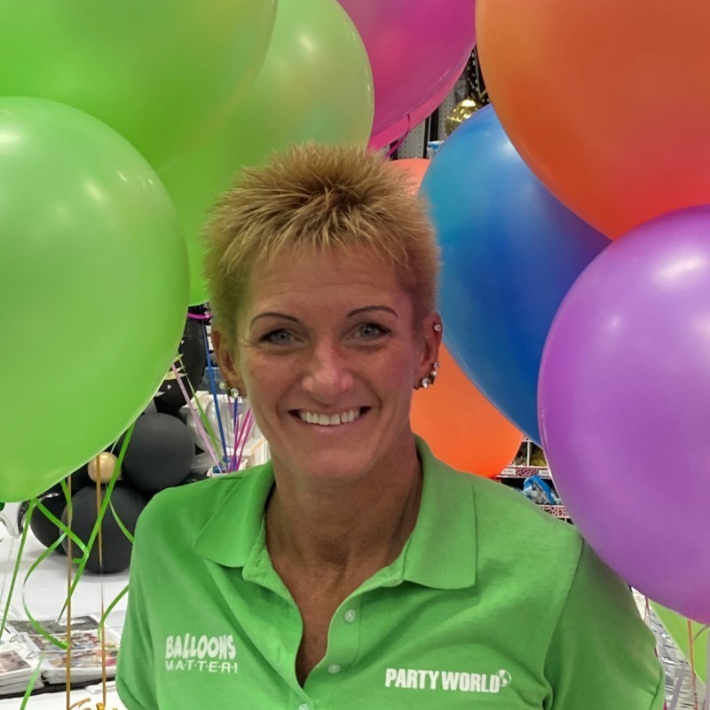 Person in a bright green Be Bold Balloons shirt surrounded by colorful balloons at an event setup
