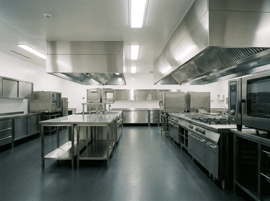 Eine hochmoderne, extrem saubere Großküche aus Edelstahl mit mehreren Arbeitsstationen, Dunstabzugshauben, professionellen Öfen, Herden und einem glänzenden, dunklen Boden.