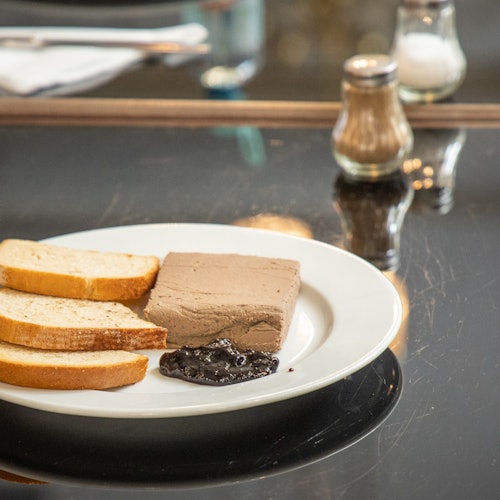 Plato con rebanadas de pan, un bloque de paté y una cucharada de mermelada sobre una mesa oscura y reflectante. Saleros y pimenteros al fondo.
