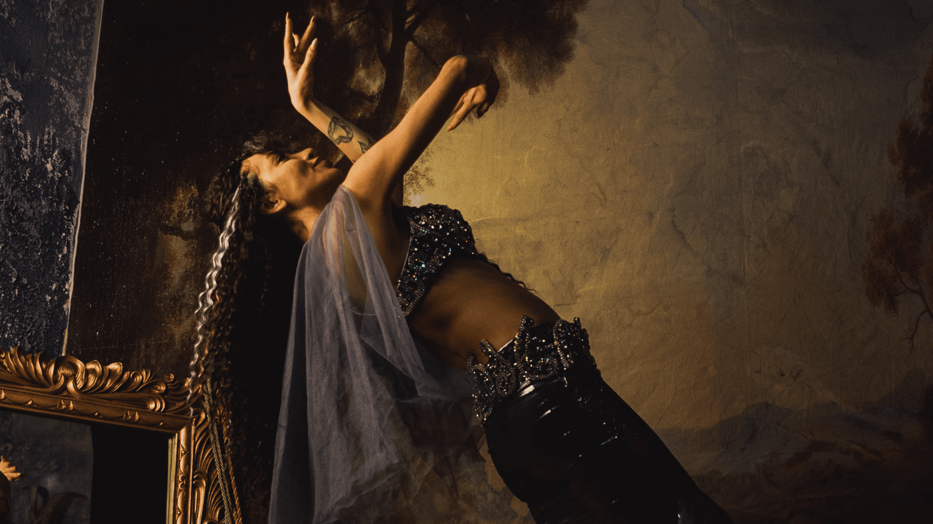 A belly dancer in a sparkling costume leans back in a fluid pose, framed by dramatic lighting and a classical painted backdrop with a golden mirror nearby.