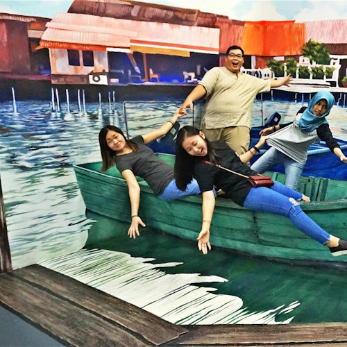 四个人在绿色船上摆着玩笑姿势，背景是一幅描绘河岸场景的壁画，有房屋和码头。