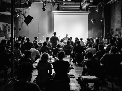 A group of people sitting in a room listening to a speaker on stage.