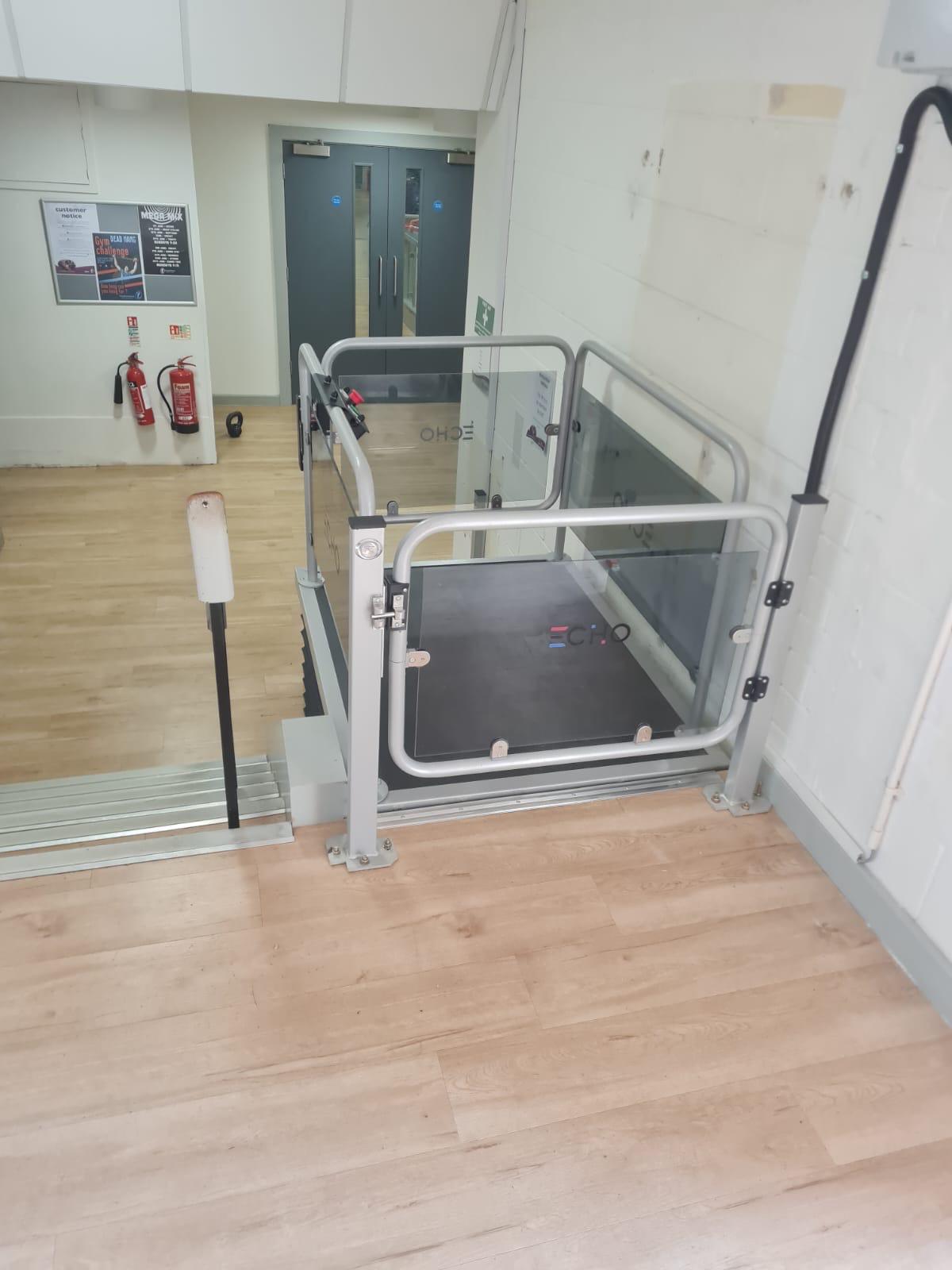 Echo platform step lift at upper level in a commercial building — open gate showing spacious non-slip platform with glass side panels, providing wheelchair access between floors in a public-facing premises
