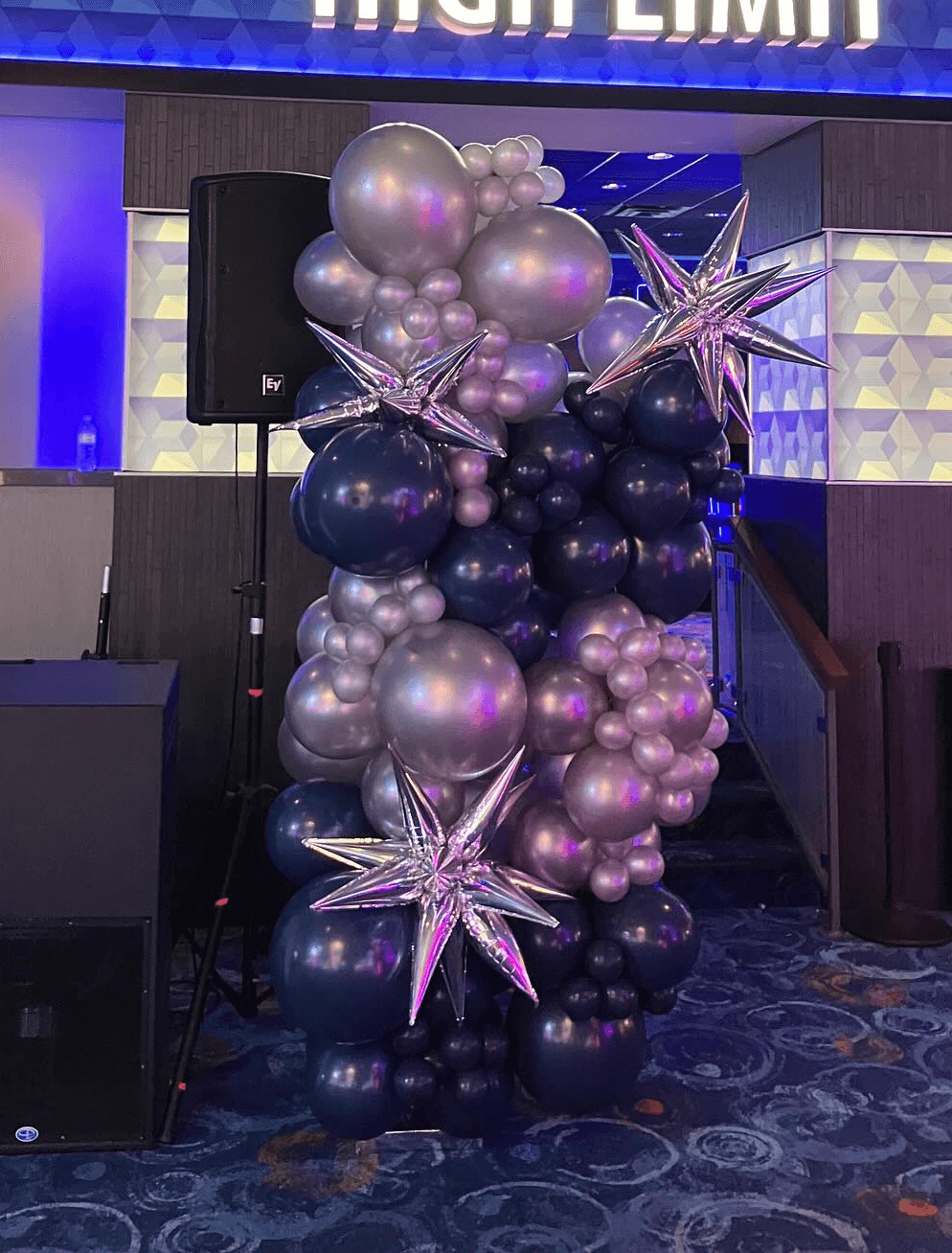 Stage decorated with purple, black, and blue balloon arch and starburst designs for an indoor event by Be Bold Balloons