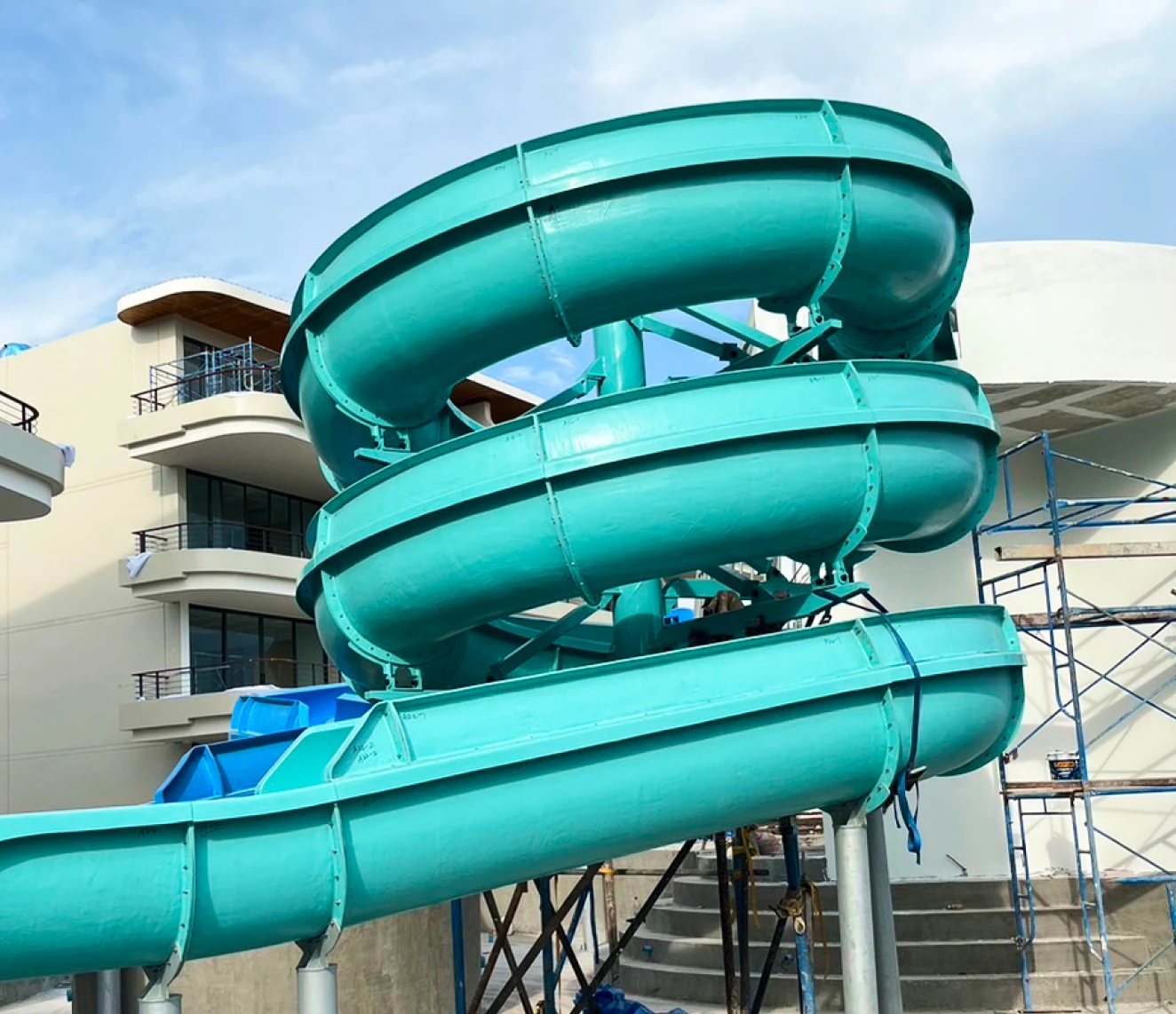 Một cầu trượt nước xoắn ốc màu xanh ngọc sôi động, nổi bật trên nền trời xanh trong, nằm gần một tòa nhà hiện đại có các ban công. Khung cảnh này truyền tải sự phấn khích và niềm vui.