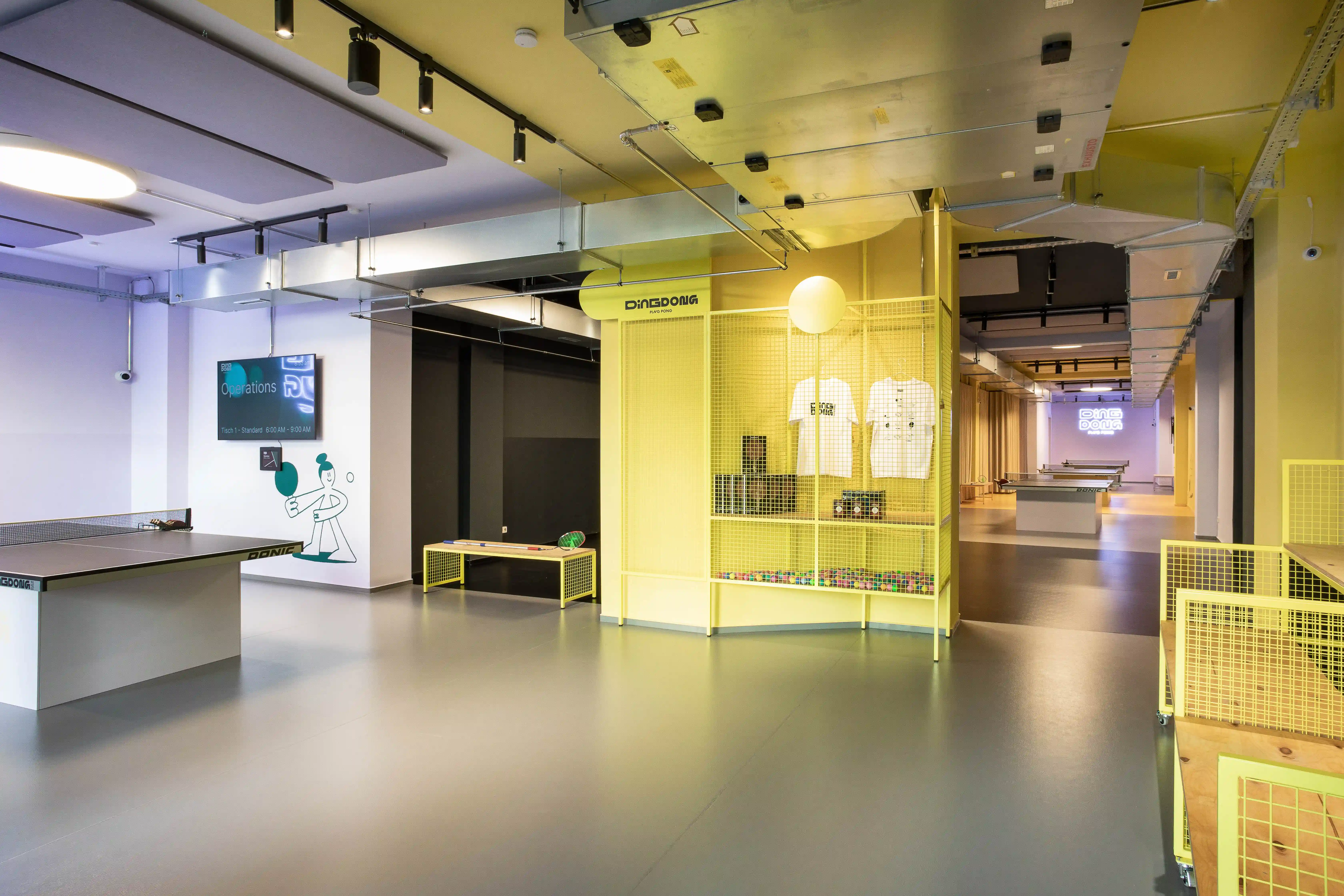 Interior of DING DONG PING PONG with a table tennis table, illustrated wall graphics, screen and the yellow mesh merchandise kiosk in the center.