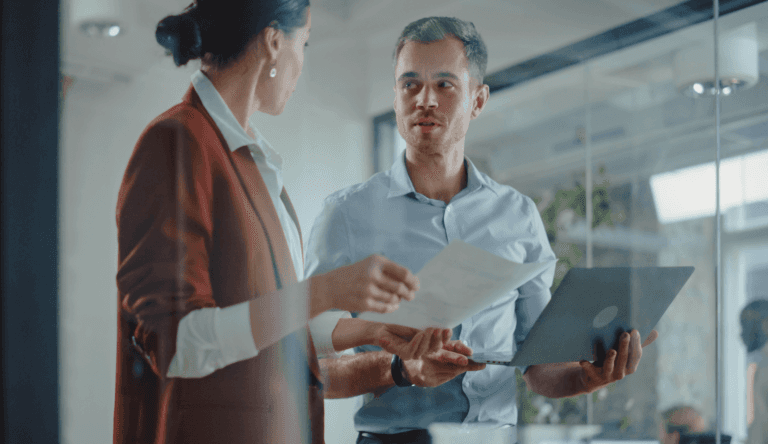 Two professionals, a woman in a red jacket and a man in a blue shirt, having a discussion in a modern office or building lobby