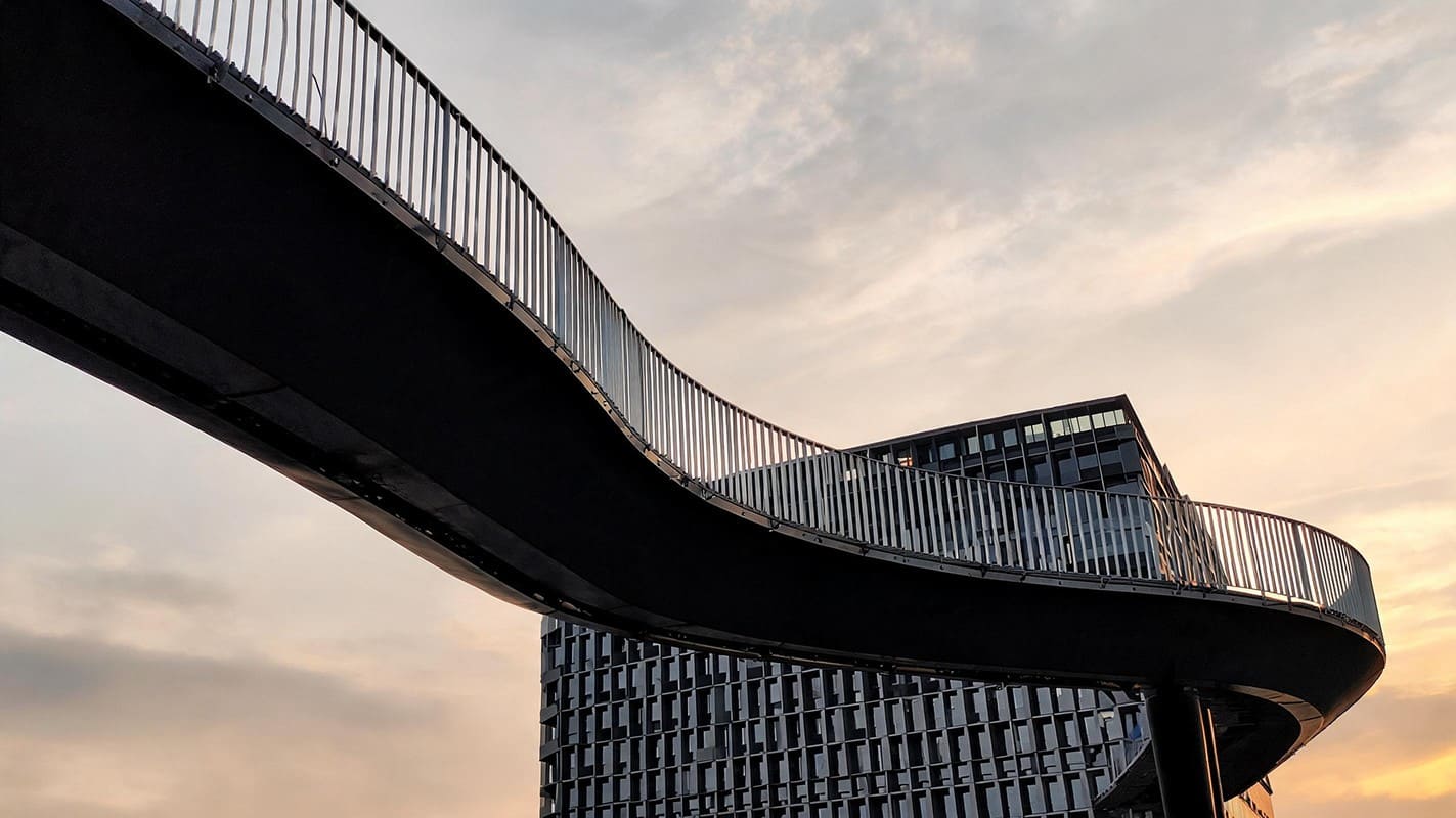 A low-angle view of a curved pedestrian overpass bridge with metal railings, sweeping across the frame in front of a modern dark gridded building during sunset.