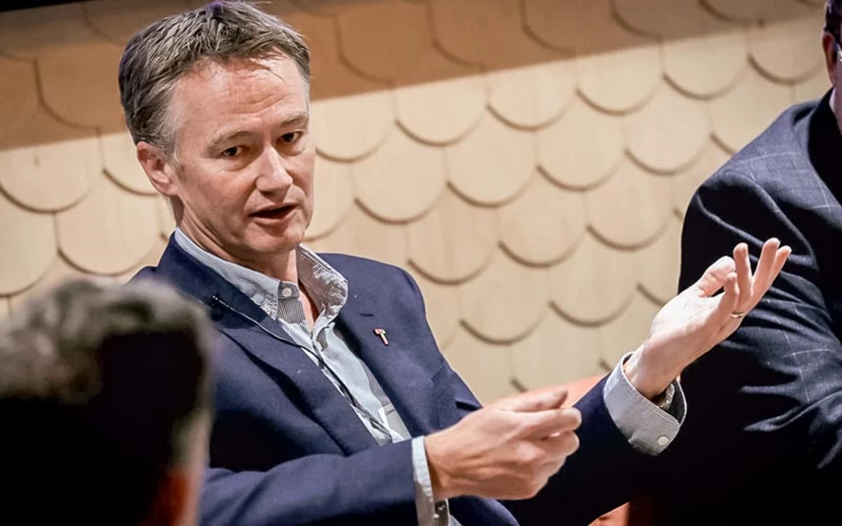 A panel speaker gestures while speaking, seated against a textured wall backdrop. Other speakers and attendees are partially visible, indicating an ongoing conversation.