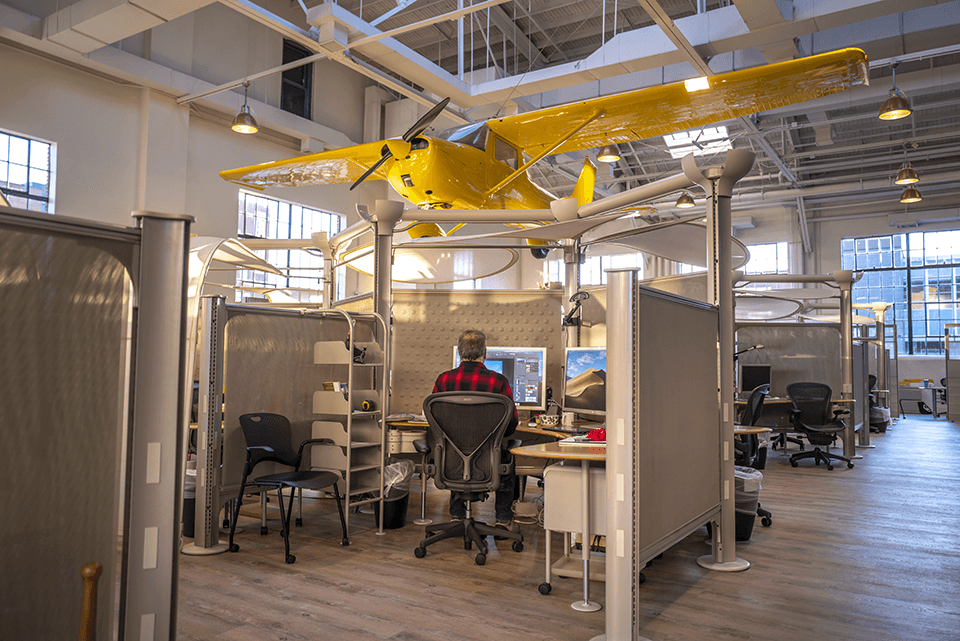 A yellow propeller plane is suspended from the ceiling inside a modern office with cubicles and large windows, reflecting the innovative spirit of this collaborative agency.